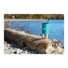 A turquoise travel mug with a metal base and straw sits on a large driftwood log. The mug is branded with meditire. Pebbles and a lake are in the background, with trees along the shore under a clear sky.