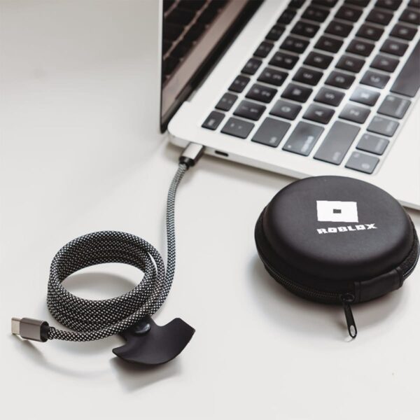 A braided cable is connected to a laptop with a partially visible keyboard. A small black case with a Roblox logo is next to it. The scene is set on a light-colored surface.