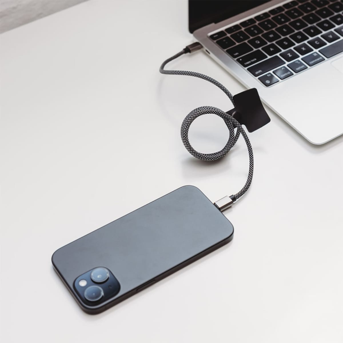 Smartphone with gray case connected to a laptop via a coiled black charging cable on a white surface. The laptop is only partially visible, showing the keyboard and charging port.