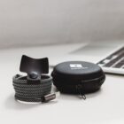 A coiled black and white braided charging cable with a small securing strap is next to a black zippered carrying case. A partially visible laptop is in the background on a white surface.
