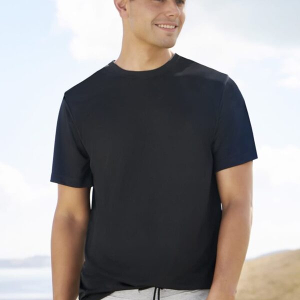 Man wearing a black T-shirt, gray sweatpants, and a dark cap smiles while standing outdoors against a backdrop of hills and a partly cloudy sky.
