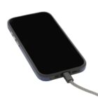 A smartphone with a black screen is charging, connected by a braided cable. The phone is in a dark-colored case, and it is positioned diagonally on a white surface.