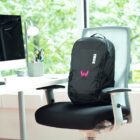 A black Thule backpack is placed on an office chair in front of a desk, which holds a computer monitor, a lamp, and a pencil holder. The window behind shows a view of lush green trees.