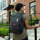 A person with short curly hair stands outside on steps, wearing a green shirt and a black backpack with a Thule logo. Theyre in front of a brick building with plants and flowers nearby.