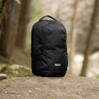 A black backpack with a logo stands on a rock in a forest setting. The background is blurred, showcasing trees and soft natural light, highlighting the outdoor, adventurous theme.