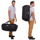A person in a light gray shirt and brown pants is holding and wearing a large black Thule duffel bag. The front view shows the person carrying the bag by its handles, while the side view shows the bag worn as a backpack.