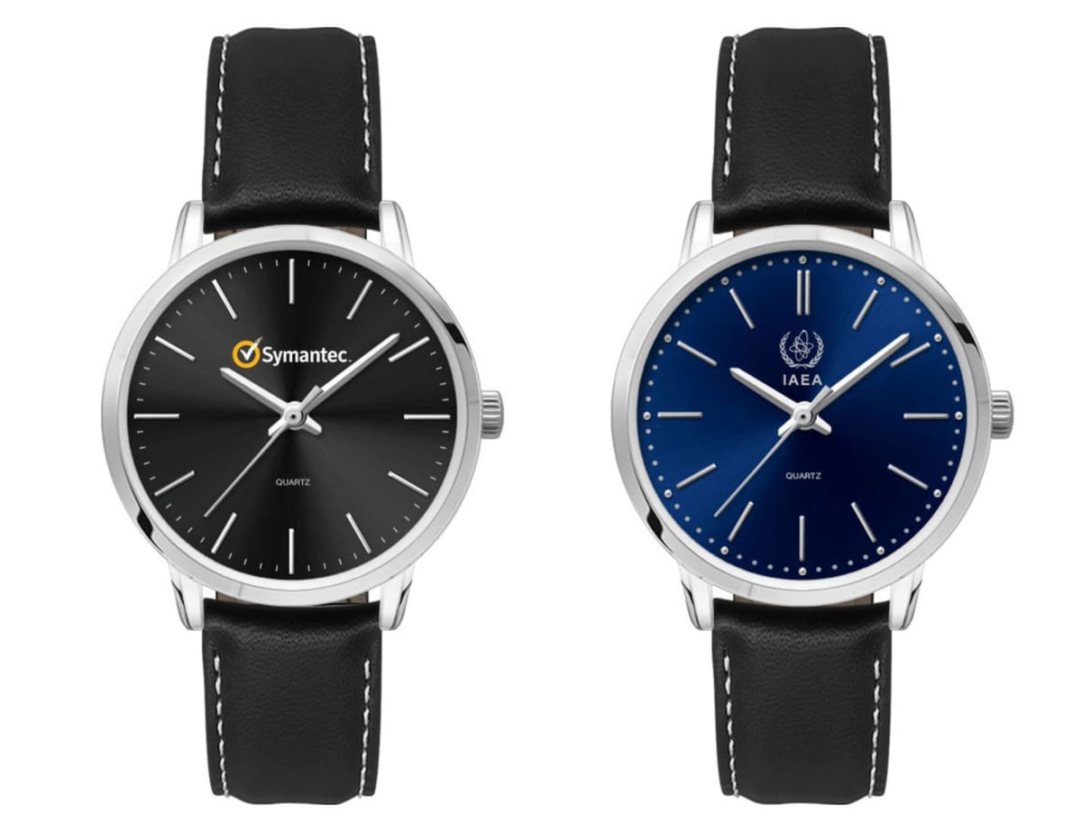 Two wristwatches with black leather straps and silver cases. The left watch has a black dial with the Symantec logo, and the right watch has a blue dial with the IAEA logo. Both have minimalist hour markers and hands.