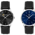 Two wristwatches with black leather straps. The left watch has a black face with the Symantec logo. The right watch has a blue face with the IAEA logo. Both have minimalist designs with silver markers and hands.