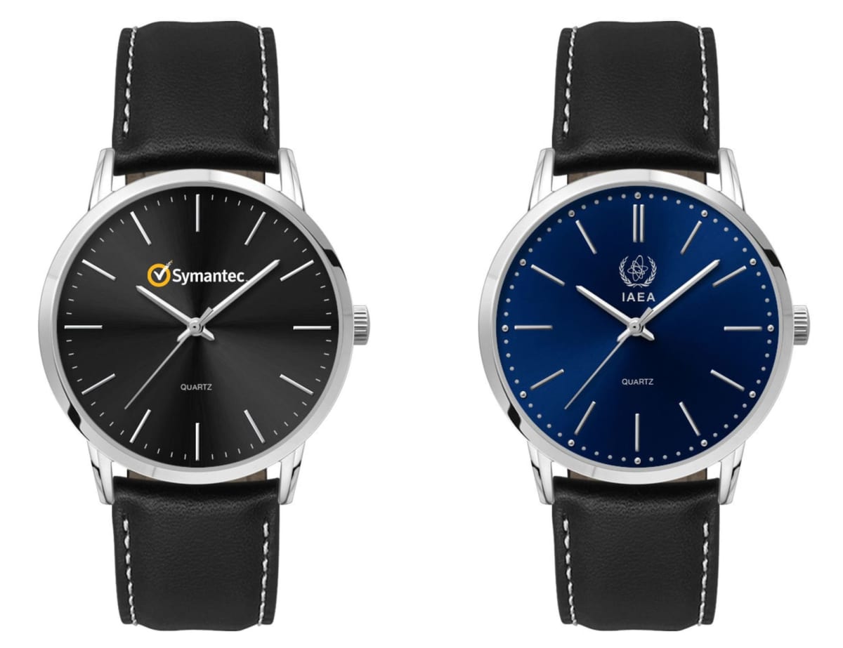 Two wristwatches with black leather straps. The left watch has a black face with the Symantec logo. The right watch has a blue face with the IAEA logo. Both have minimalist designs with silver markers and hands.