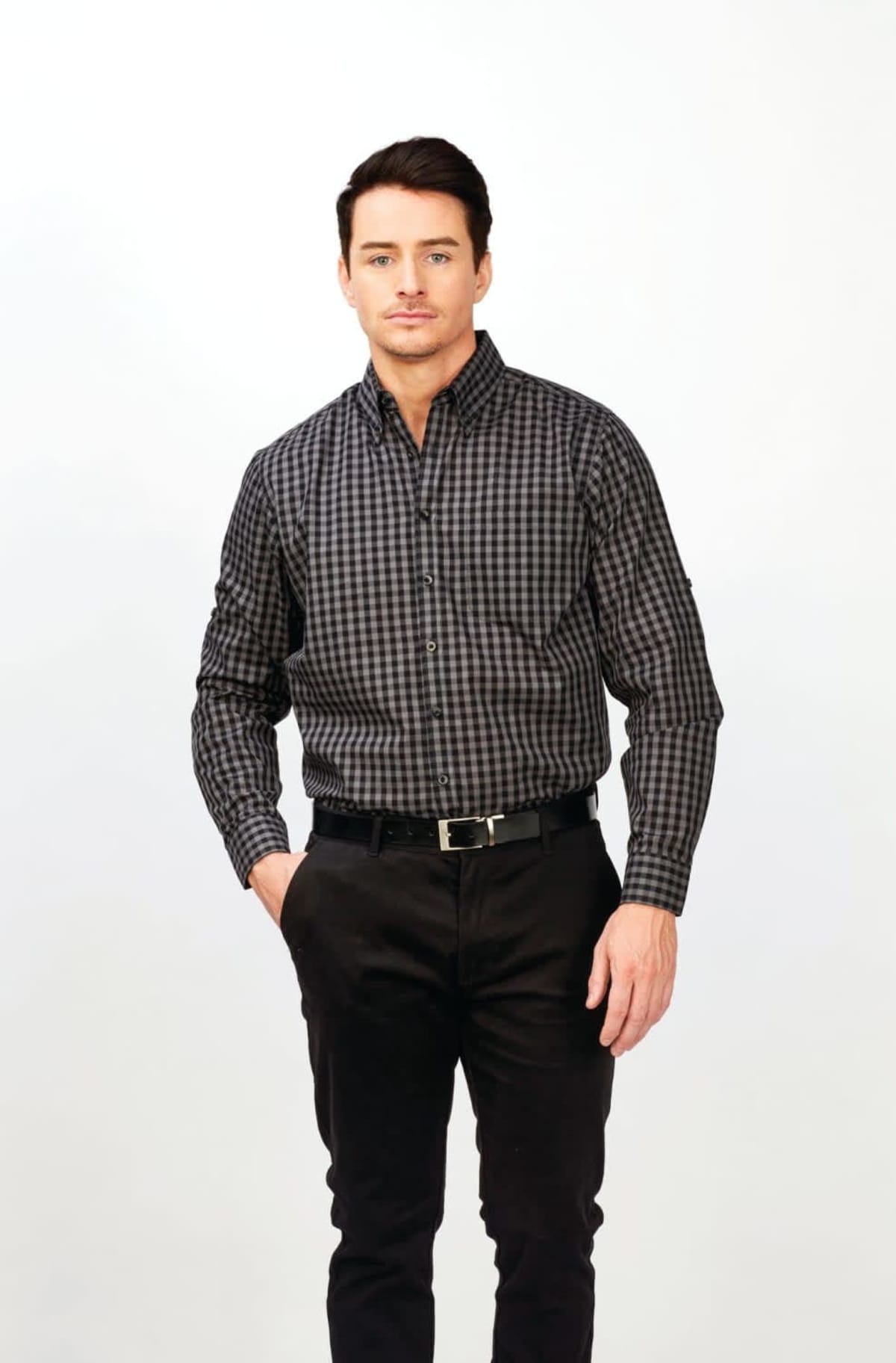 A man with dark hair is wearing a black and gray checkered long-sleeve shirt and black pants. His left hand rests in his pocket, and he stands against a light gray background.