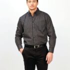 A person standing against a white background, wearing a black and gray checkered button-up shirt and black pants. They have their left hand in their pants pocket and are looking at the camera.