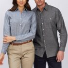A woman and a man standing side by side against a plain background. The woman wears a light blue button-up shirt and beige pants. The man wears a dark gray button-up shirt and navy shorts. They both have neutral expressions.