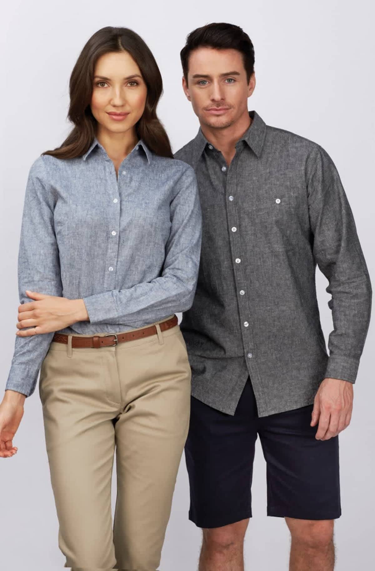 A woman and a man standing side by side against a plain background. The woman wears a light blue button-up shirt and beige pants. The man wears a dark gray button-up shirt and navy shorts. They both have neutral expressions.