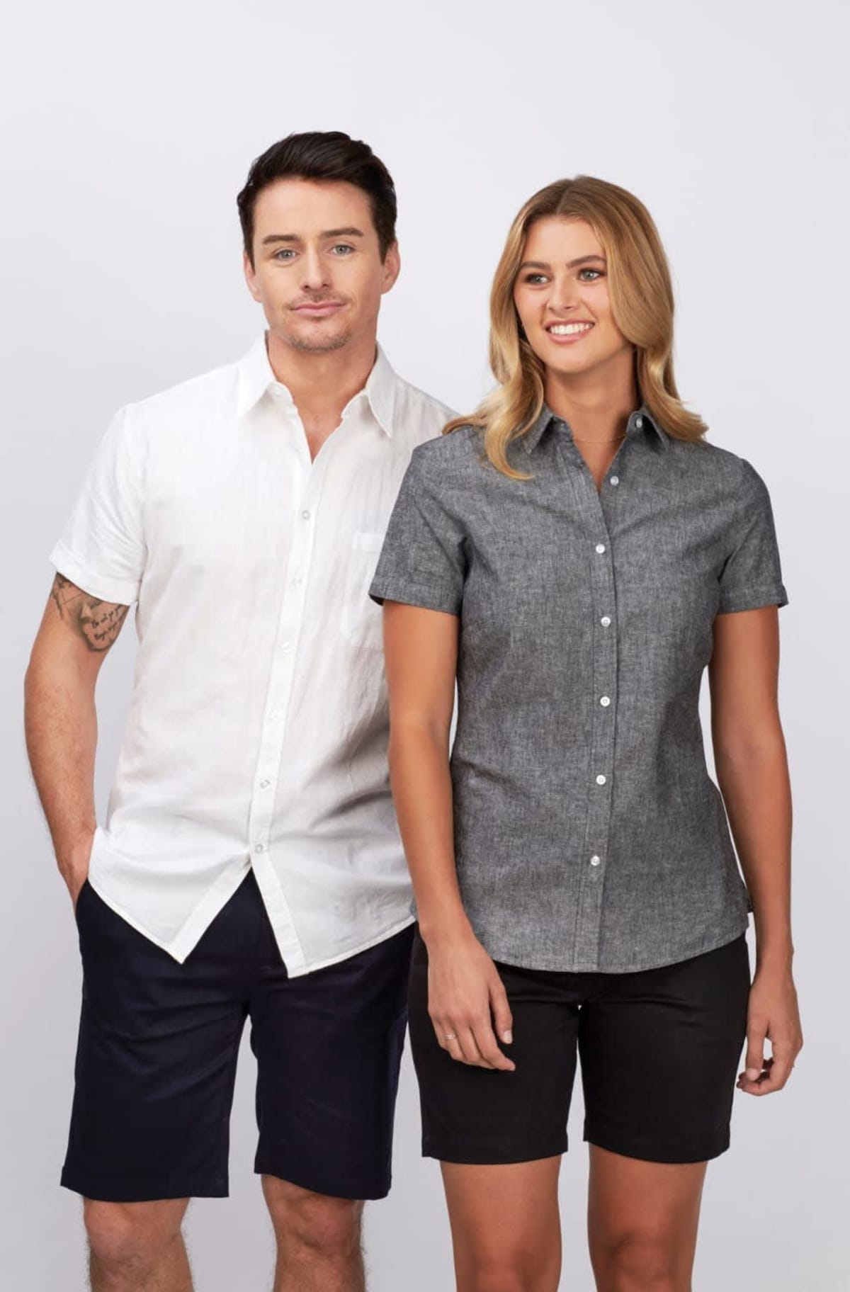 A man in a white short-sleeve shirt and black shorts stands with a woman in a dark short-sleeve shirt and black shorts. They are both smiling against a plain background.
