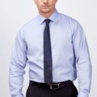 A man wearing a light blue dress shirt, dark tie, and black pants stands against a plain white background, looking directly at the camera with a neutral expression.