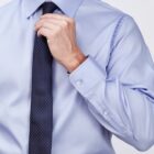 A person adjusts their dark, dotted tie while wearing a light blue dress shirt. The shirt has buttoned cuffs and is tucked into black trousers. The background is a plain, light color.