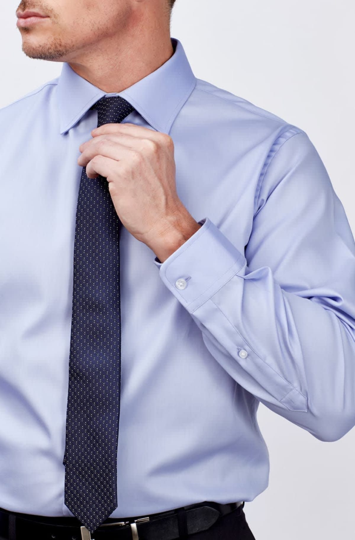 A person adjusts their dark, dotted tie while wearing a light blue dress shirt. The shirt has buttoned cuffs and is tucked into black trousers. The background is a plain, light color.