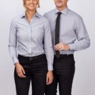 Two individuals stand side by side against a plain background. Both are wearing light gray long-sleeve button-up shirts and black pants. The person on the right also wears a black tie. They are smiling and have a relaxed posture.