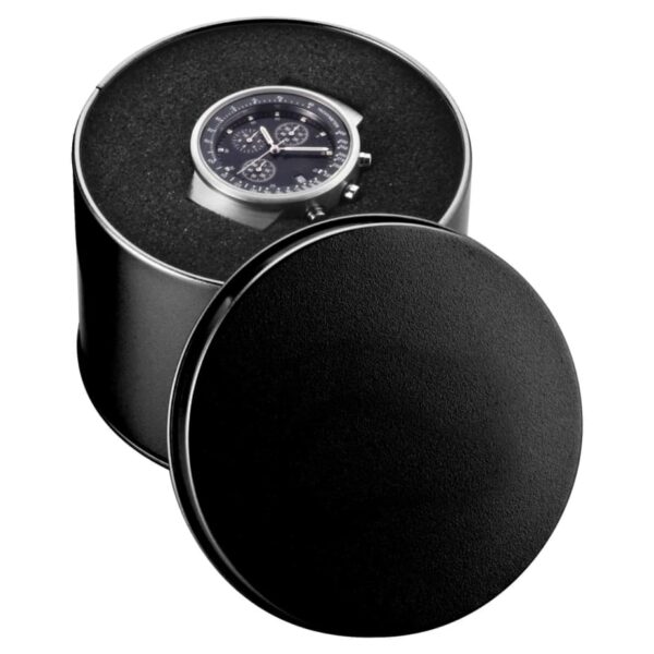 A wristwatch with a black dial and silver case is displayed in a round, black-leather case. The lid is partially open, revealing the watch inside. The case has a soft, textured interior.
