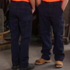 Two individuals in navy blue cargo pants and bright orange shirts stand facing each other in a workshop setting. Their faces are not visible. One wears black boots, and the other wears brown work boots. Various workshop equipment is partially visible in the background.