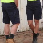 Two people in bright yellow shirts and dark cargo shorts stand on a brick sidewalk. One has brown boots with socks, and the other wears black boots with black socks. They face each other, with a white wall in the background.