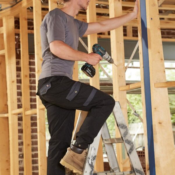 A person with long hair tied back is standing on a ladder, holding a power drill, working on wooden framing inside a construction site. They are wearing a gray shirt, black work pants, and boots. Sunlight filters through the structure.