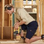 A person wearing ear protection and a beige shirt kneels while operating a yellow power drill on the wooden frame of a building under construction. They are working in a partially built structure with exposed wooden beams.