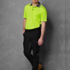 A man stands against a gray textured background wearing a bright green polo shirt, black pants, and tan work boots. His hands are in his pockets, and he looks at the camera with a slight smile.