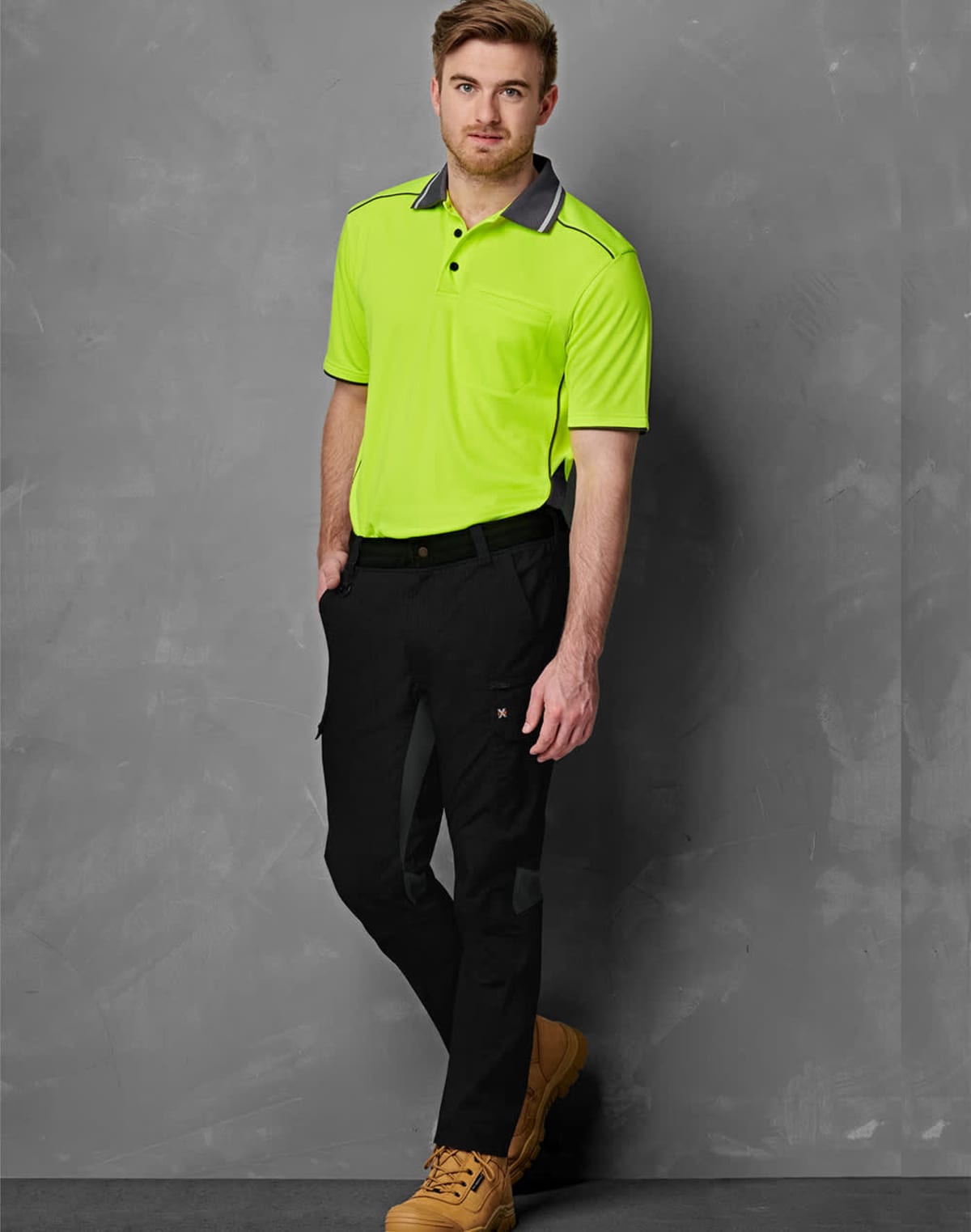 A man stands against a gray textured background wearing a bright green polo shirt, black pants, and tan work boots. His hands are in his pockets, and he looks at the camera with a slight smile.