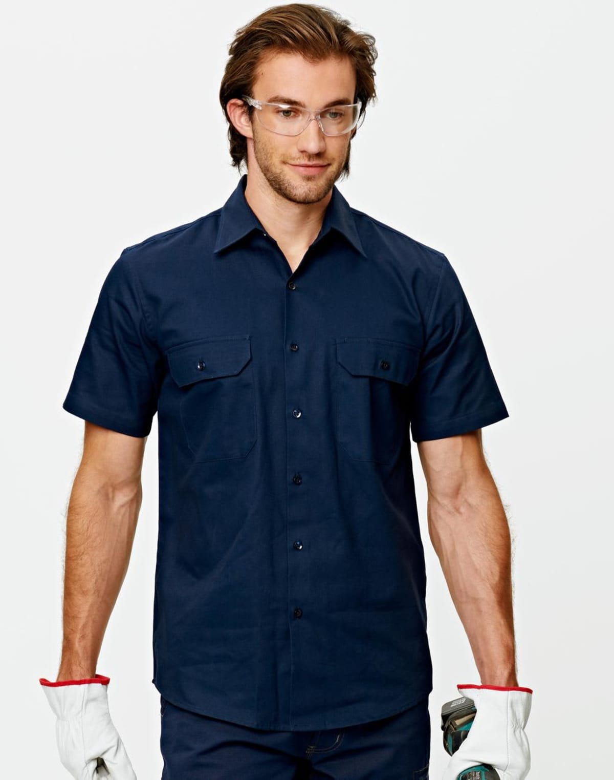 A person wearing safety glasses and a navy blue short-sleeve button-up shirt holds work gloves. They have a neutral expression and are standing against a plain background.