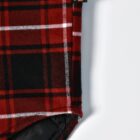 Close-up of a red and black plaid fabric with white lines, featuring a small black tag with orange and white design. The fabric is folded with a visible black lining underneath on a white background.
