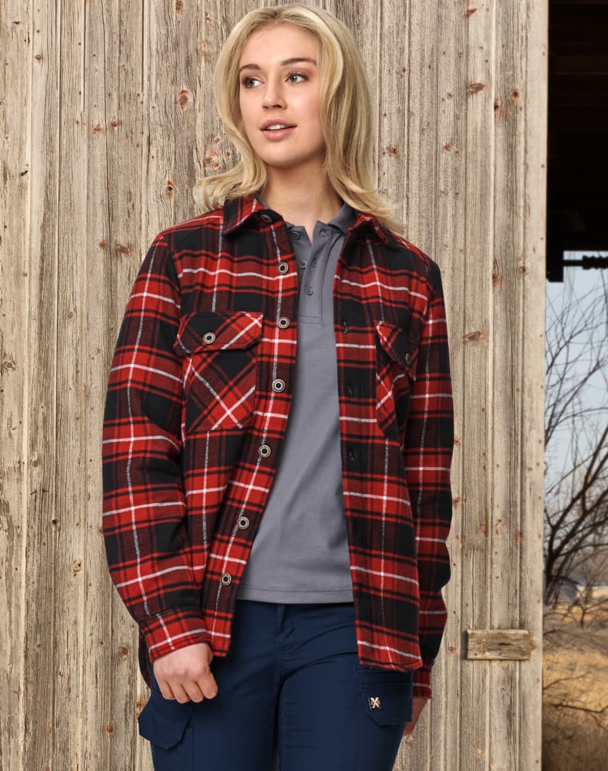 A person with long blonde hair wears a red and black plaid shirt over a gray top and blue pants, standing against a weathered wooden wall with a glimpse of a rural landscape in the background.