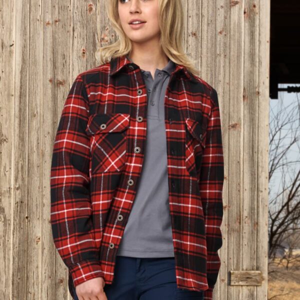 A person with long blonde hair stands casually in front of a weathered wooden wall. They are wearing a red and black plaid shirt over a gray top, paired with blue pants. The background includes dry grass and bare tree branches.