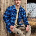 A person with long blonde hair wearing a blue plaid shirt and khaki pants sits on a wooden structure. The background features a wooden building and a field with sparse trees.
