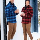 Two people stand outdoors in the snow, wearing blue and red plaid jackets, brown pants, and boots. They are smiling and holding mugs, with a stack of firewood nearby and a snow-covered landscape in the background.