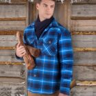 A man in a blue plaid shirt and scarf holds firewood and an ax, standing in front of a wooden wall with snow on the roof. He gazes to the side, creating a wintery, rustic atmosphere.