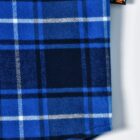 Close-up of a blue plaid fabric with various shades of blue and intersecting white lines. A small black tag with orange text is attached to the edge of the fabric.