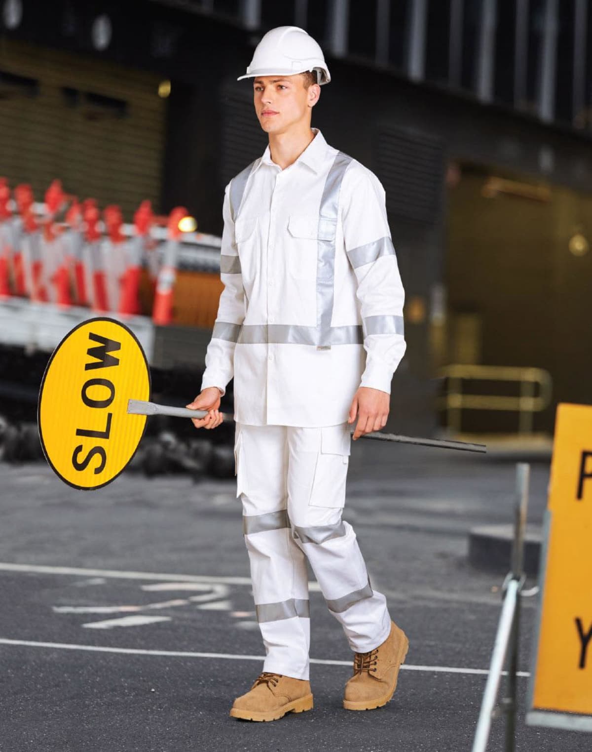 A construction worker wearing a white uniform, helmet, and reflective strips holds a yellow SLOW sign in a street setting. Orange cones are visible in the background, along with a partially seen structure.