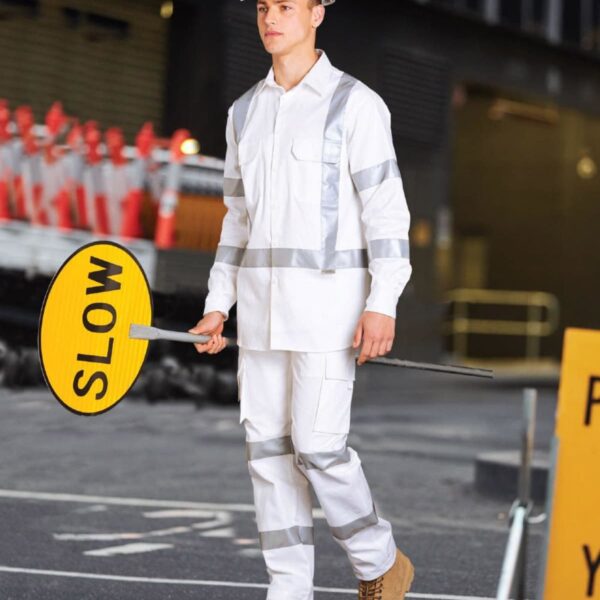 A construction worker in a white uniform and hard hat holds a yellow SLOW sign. He stands on a paved area with traffic cones and a dark building in the background.
