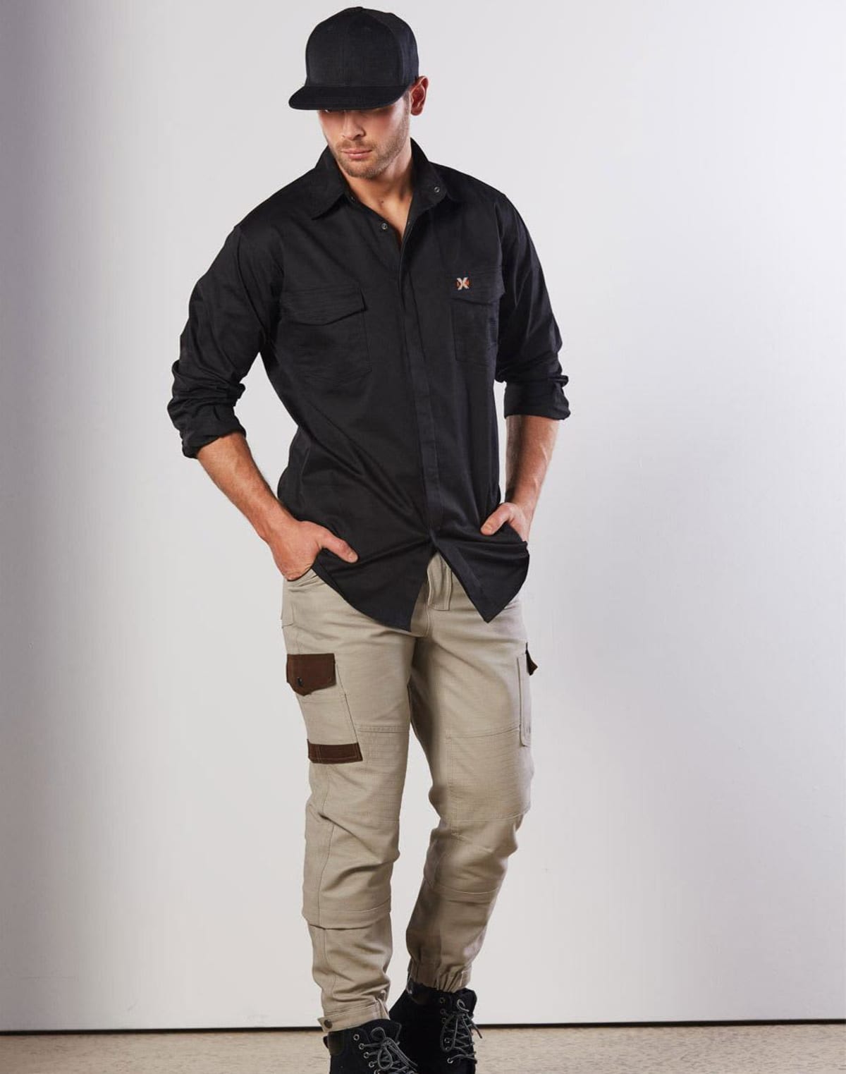 A man wearing a black cap, a black long-sleeve shirt, and beige cargo pants with pockets poses against a white background. He is looking down with his hands in his pockets.