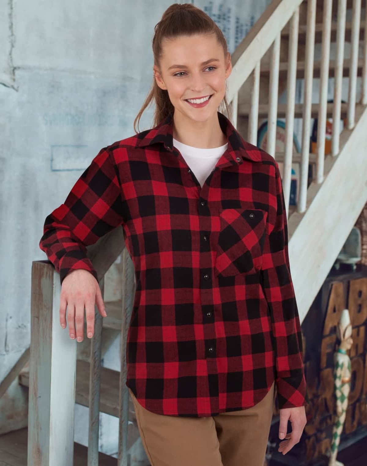 A person with long hair stands indoors, smiling, and wearing a red and black plaid shirt over a white top. They have one hand resting on a wooden railing. Stairs and a painted wall are visible in the background.