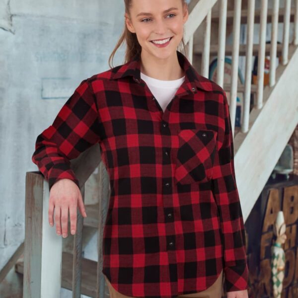 A person wearing a red and black plaid shirt and khaki pants stands indoors, smiling and leaning on a railing. They have long, light brown hair tied back and are in front of a set of stairs.