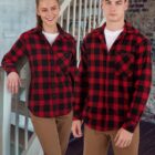A woman and a man stand side by side on a staircase, both wearing red and black plaid shirts with white undershirts and brown pants. The background features a mix of brick and painted walls.