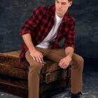 A man wearing a red and black plaid shirt, white t-shirt, and tan pants sits on a wooden crate. He looks down, with a thoughtful expression. He has short dark hair and wears dark sneakers. The background is a textured dark gray.