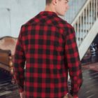 A person wearing a red and black plaid shirt stands in a room with wooden floors and a staircase. The person is turned away, looking slightly to the side. In the background, theres a brown sofa.
