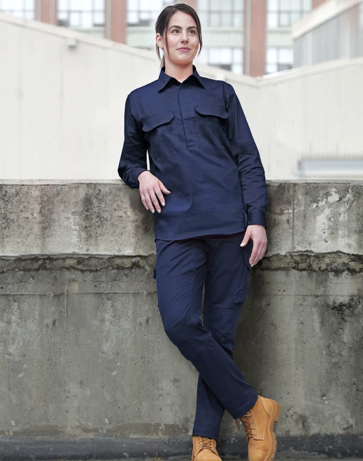 Person in navy work attire and tan boots, leaning casually against a concrete wall. The setting appears to be an urban environment with buildings in the background.