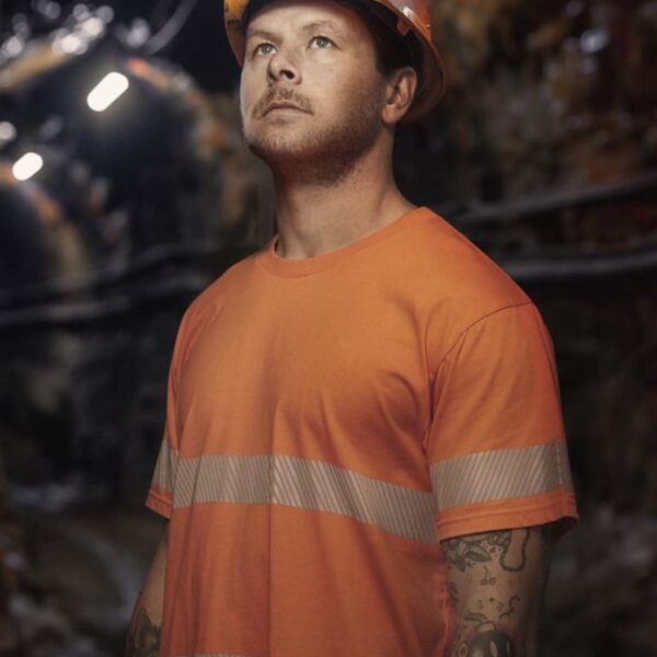 A person wearing an orange safety shirt and helmet with a headlamp stands in an underground mine tunnel, looking upwards. The background is dimly lit, highlighting rocky walls and electrical cabling. They have tattoos on their arms.