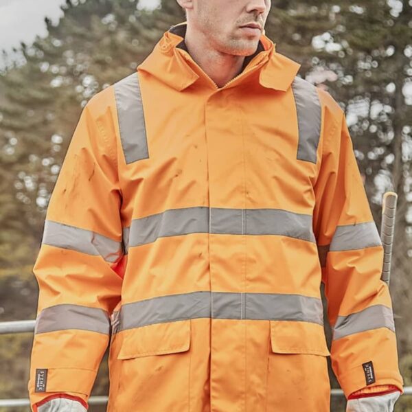 A person wearing an orange high-visibility jacket and a white hard hat stands outdoors at a construction site, with trees in the background. They have protective gloves and are looking to the side.