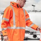 A person wearing an orange high-visibility jacket with reflective stripes and a white hard hat stands outdoors near traffic cones. The background shows a cloudy sky and industrial equipment.
