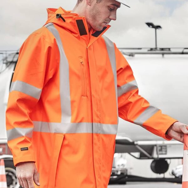 A person wearing an orange high-visibility jacket with reflective stripes and a white hard hat stands outdoors near traffic cones. The background shows a cloudy sky and industrial equipment.